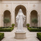 White marble religious woman statue displayed in a church courtyard, featuring flowing robes, open hands, detailed carving, and a peaceful sacred architectural setting.
