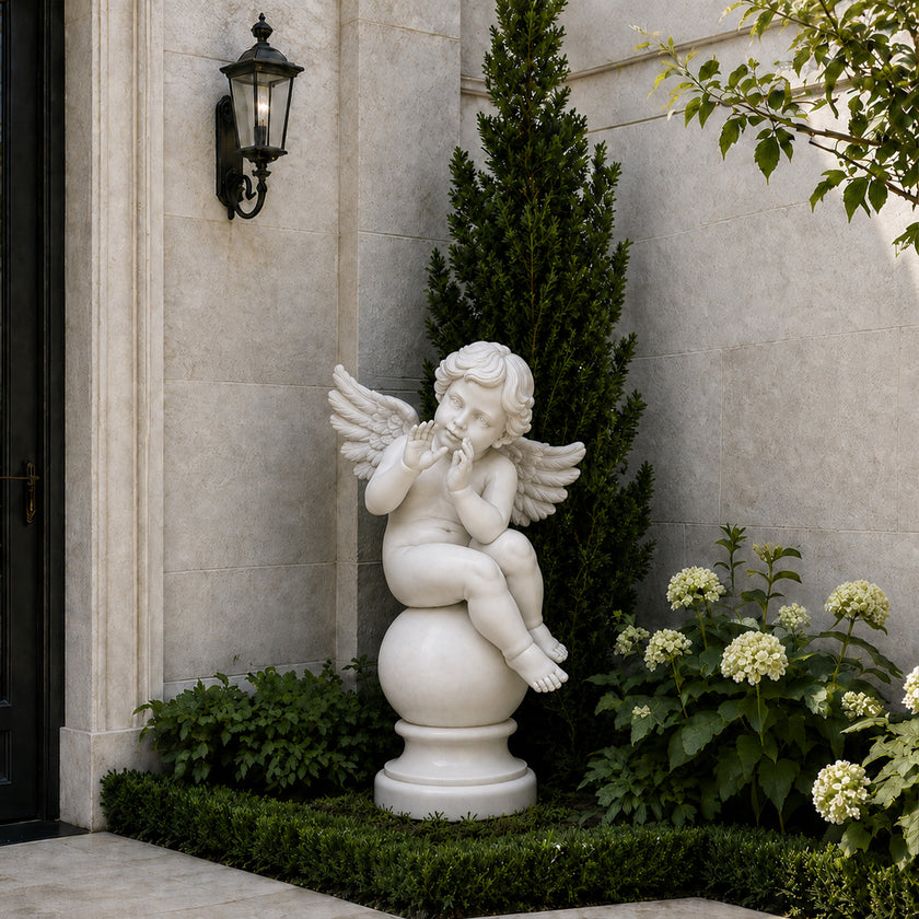 A white marble-style cherub garden statue seated on a round pedestal, placed in a landscaped garden corner with green shrubs, white flowers, a tall evergreen tree, and a beige stone wall with a black wall sconce.