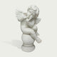 A white marble cherub figurine sitting on a round pedestal, with small wings and a thoughtful expression, photographed against a light gray background.