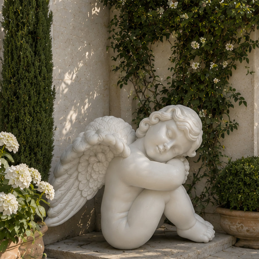 White marble sleeping angel statue with folded arms, delicate wings and serene expression displayed in an elegant garden setting.