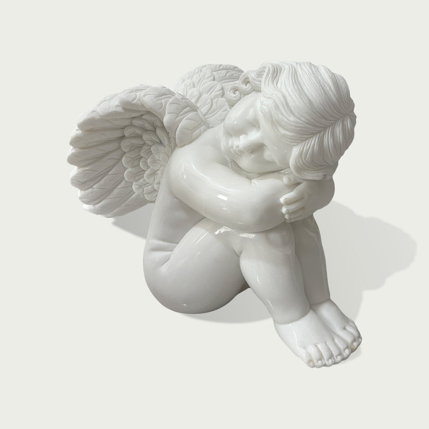 Classical white marble cherub sculpture featuring carved wings, curled hair and a peaceful resting pose for indoor or outdoor decor.