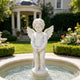White marble cherub angel fountain statue displayed in a garden setting, with detailed wings, standing pose, and water feature design for outdoor decoration.