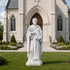 White marble saint statue wearing flowing robes, holding a book and lily flowers, displayed in front of a historic church entrance with classical architectural details.