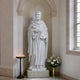 White marble religious statue of a saint with detailed robe folds, holding a sacred book and flowers, placed inside an elegant church interior with soft lighting.