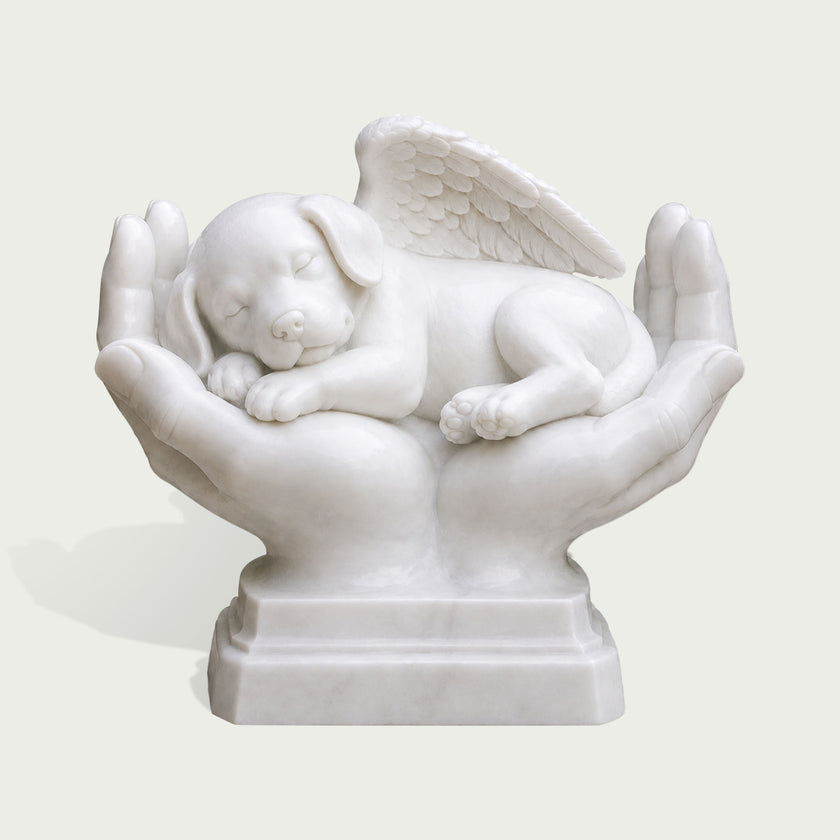 White marble pet memorial sculpture featuring a peaceful sleeping puppy with a detailed feathered angel wing, cradled in two caring hands on a carved pedestal base.