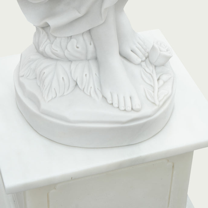 Close-up of the white marble woman statue base showing bare feet, carved leaves and flowers, round plinth, and square pedestal with a smooth finish.