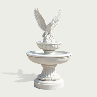 White marble eagle fountain sculpture shown on a clean neutral background, featuring an eagle with fully spread wings perched on a rocky formation above two tiered circular basins, with carved petal details, a fluted pedestal, and a round marble base.