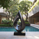 Stainless Steel Abstract sculpture by a pool with modern building and greenery in the background