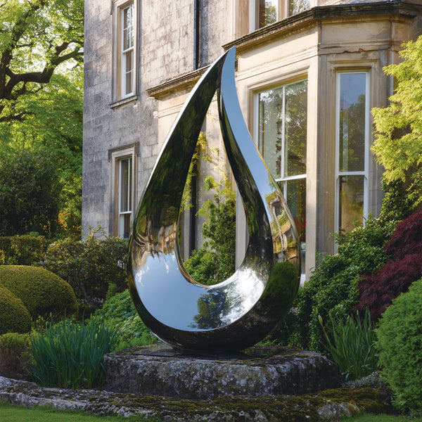 Large stainless steel hollow teardrop sculpture in a garden setting with a classical building in the background