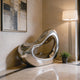 Modern stainless steel silver liquid cloud sculpture in a stylish interior setting with a lamp and plant.
