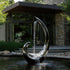 Abstract mirror-polished stainless steel teapot sculpture with a flowing water feature in front of a house.