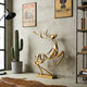 Openwork Golden Stag Abstract Stainless Steel Sculpture displayed in a styled room setting with a concrete wall, framed posters, a potted cactus, a black floor lamp, and a bookshelf.