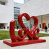 Charming LOVE Lettering Stainless Steel Floor Sculpture  in front of a modern white building with glass windows and a few people walking in the background.