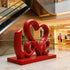 Charming LOVE Lettering Stainless Steel Floor Sculpture on a matching red base in a modern shopping mall atrium with escalators and store fixtures in the background.