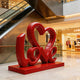 Charming LOVE Lettering Stainless Steel Floor Sculpture on a matching red base in a modern shopping mall atrium with escalators and store fixtures in the background.