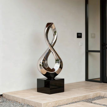 Gleaming Infinity Abstract Stainless Steel Sculpture on a glossy black rectangular base, displayed on a stone terrace in front of a minimalist building.