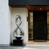 Gleaming Infinity Abstract Stainless Steel Sculpture on a black rectangular pedestal in front of a contemporary building entrance with a black door and white wall.