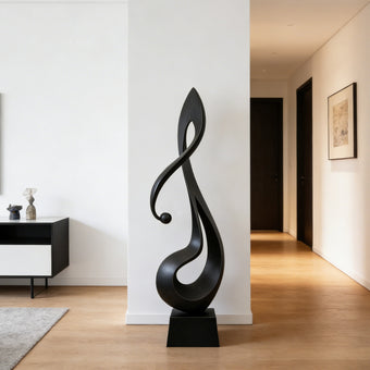 Lyrical Musical Note Black Stainless Steel Sculpture on a rectangular black base, set outdoors on a light stone patio with green grass and a minimalist concrete building in the background.
