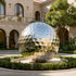 Mirror Polished Golf Ball Contemporary Stainless Steel Sculptures sits outdoors in a landscaped courtyard in front of a beige villa with arched windows and a tree.
