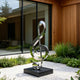 Modern Musical Note Outdoor Stainless Steel Sculpture on a black rectangular base, set in a modern landscaped garden with white gravel, green plants, and a contemporary wooden house in the background.