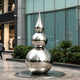 Striking Teardrop Tower Outdoor Stainless Steel Sculpture with a stacked spherical and teardrop silhouette stands on a circular base in an outdoor plaza in front of a modern glass building.