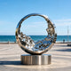 Mesmerizing Tidal Circle Abstract Stainless Steel Sculpture on a cylindrical base, photographed outdoors on a paved plaza with the ocean and blue sky in the background.