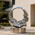 Mesmerizing Tidal Circle Abstract Stainless Steel Sculpture of a ring with a textured, fluid-like interior, mounted on a cylindrical base in an outdoor plaza with a modern building in the background.
