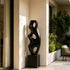 Abstract Large Voids Stainless Steel Sculpture with three large oval cutouts stands on a square black pedestal in a sunlit modern patio corridor with beige walls and green plants.