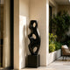 Abstract Large Voids Stainless Steel Sculpture with three large oval cutouts stands on a square black pedestal in a sunlit modern patio corridor with beige walls and green plants.