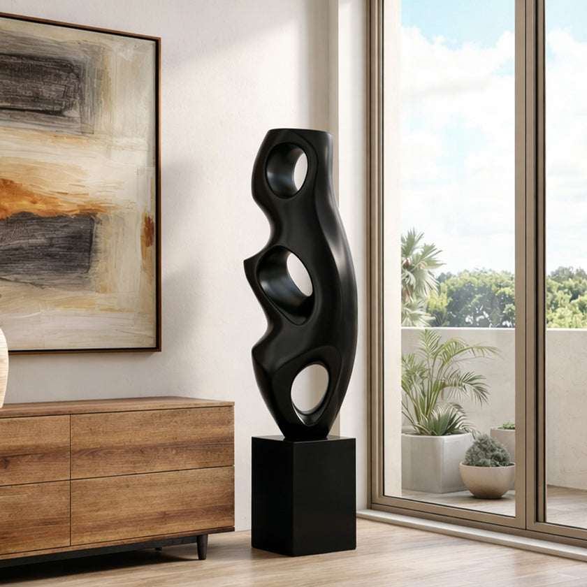 Abstract Large Voids Stainless Steel Sculpture with organic wavy curves and circular cutouts, displayed on a black pedestal in a bright living room next to a wooden sideboard and large windows.
