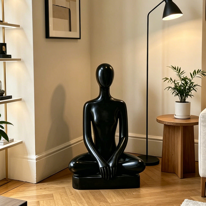 Peaceful Meditating Seated Figure Stainless Steel Indoor Sculpture displayed in a modern living room corner with a wooden floor, a black floor lamp, and a side table with a potted plant.