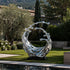 Dynamic Splashing Wave Mirror Stainless Steel Sculpture Large Outdoor Statuaryresembling a splashing or flowing wave, displayed outdoors on a black base in a landscaped garden with grass, trees, and a pool in the foreground.
