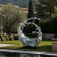 Dynamic Splashing Wave Mirror Stainless Steel Sculpture Large Outdoor Statuaryresembling a splashing or flowing wave, displayed outdoors on a black base in a landscaped garden with grass, trees, and a pool in the foreground.