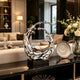 Dynamic Splashing Wave Mirror Stainless Steel Sculpture Large Outdoor Statuary on a black rectangular base, displayed on a dark console table in a modern, upscale living room setting next to a floral arrangement and decorative plate.