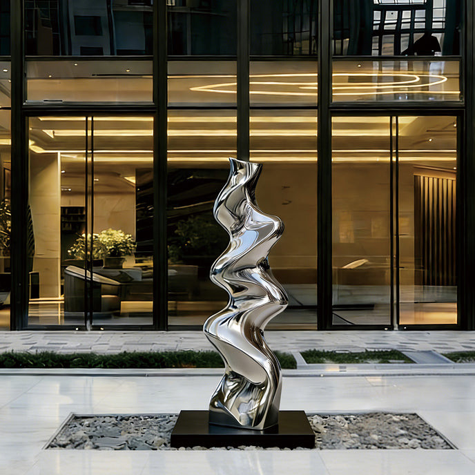 Vertical Liquid Ripple Abstract Stainless Steel Sculpture with a wavy, flowing silhouette in polished silver, displayed on a black rectangular base in front of a contemporary glass building lobby.
