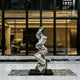 Vertical Liquid Ripple Abstract Stainless Steel Sculpture with a wavy, flowing silhouette in polished silver, displayed on a black rectangular base in front of a contemporary glass building lobby.