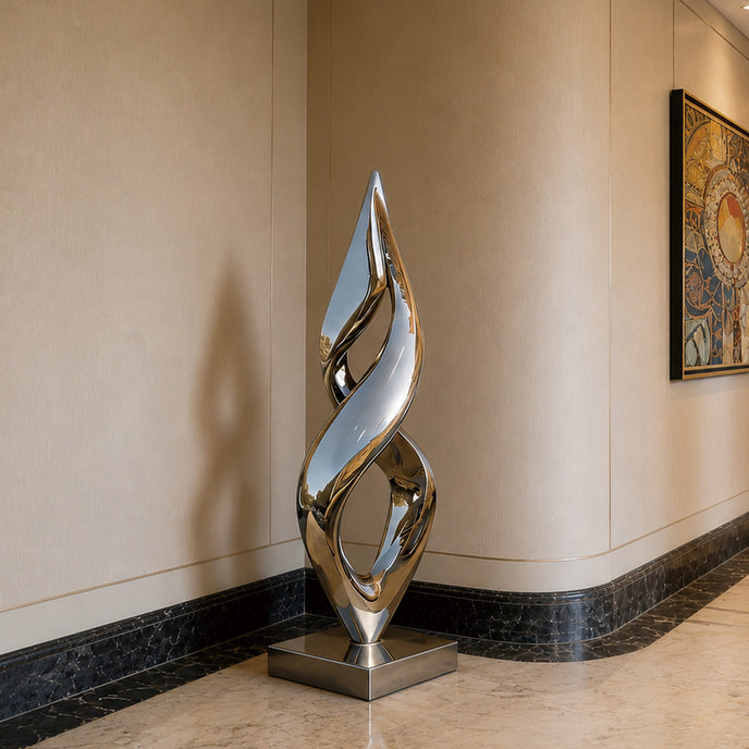 A modern abstract stainless steel sculpture with a polished reflective finish, standing on a square base in a hallway with beige walls and dark marble trim.
