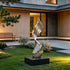 A modern abstract stainless steel sculpture with a swirling, ribbed design stands on a black rectangular base in a landscaped garden courtyard in front of a contemporary house.