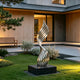 A modern abstract stainless steel sculpture with a swirling, ribbed design stands on a black rectangular base in a landscaped garden courtyard in front of a contemporary house.