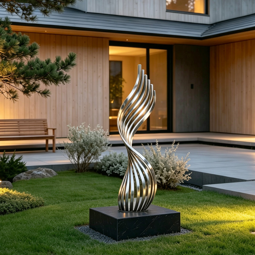 A modern abstract stainless steel sculpture with a swirling, ribbed design stands on a black rectangular base in a landscaped garden courtyard in front of a contemporary house.