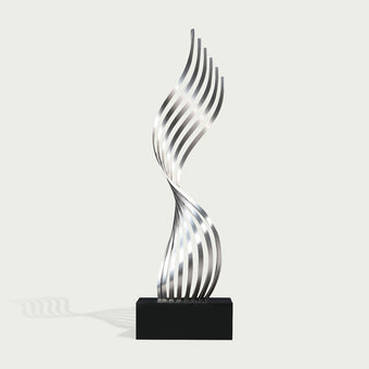 A modern abstract sculpture featuring a metallic silver, wavy, ribbon-like form rising from a solid black rectangular base, set against a light off-white background.