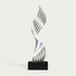 A modern abstract sculpture featuring a metallic silver, wavy, ribbon-like form rising from a solid black rectangular base, set against a light off-white background.