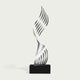 A modern abstract sculpture featuring a metallic silver, wavy, ribbon-like form rising from a solid black rectangular base, set against a light off-white background.