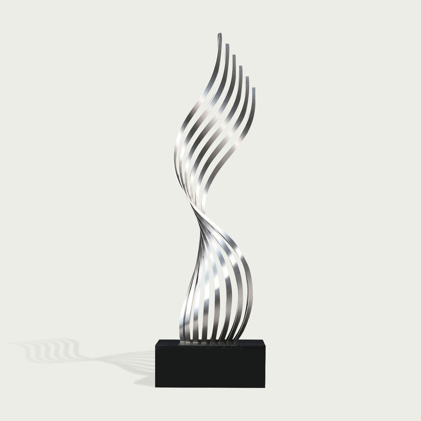 A modern abstract sculpture featuring a metallic silver, wavy, ribbon-like form rising from a solid black rectangular base, set against a light off-white background.