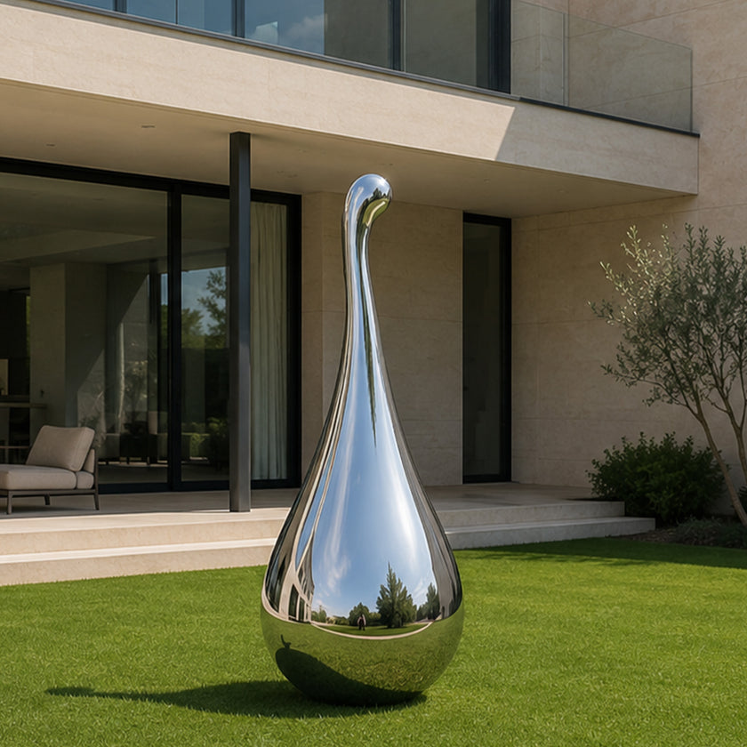 A large polished chrome sculpture shaped like a teardrop or pear stands on a green lawn in front of a modern house with glass walls and beige stone exterior.