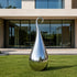 A large, teardrop-shaped polished chrome sculpture stands on a green lawn in front of a modern house with large glass windows.