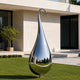 A large teardrop-shaped polished stainless steel sculpture stands on a green lawn in front of a modern beige building with dark glass windows.