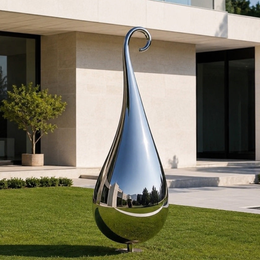 A large teardrop-shaped polished stainless steel sculpture stands on a green lawn in front of a modern beige building with dark glass windows.
