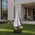 Side-Hook Curved Water Drop Mirror Polished Stainless Steel Sculpture on a lawn in front of a modern villa