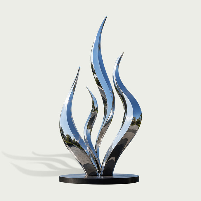 A modern abstract flame-shaped sculpture made of highly polished reflective metal, mounted on a round black base against a light gray background.